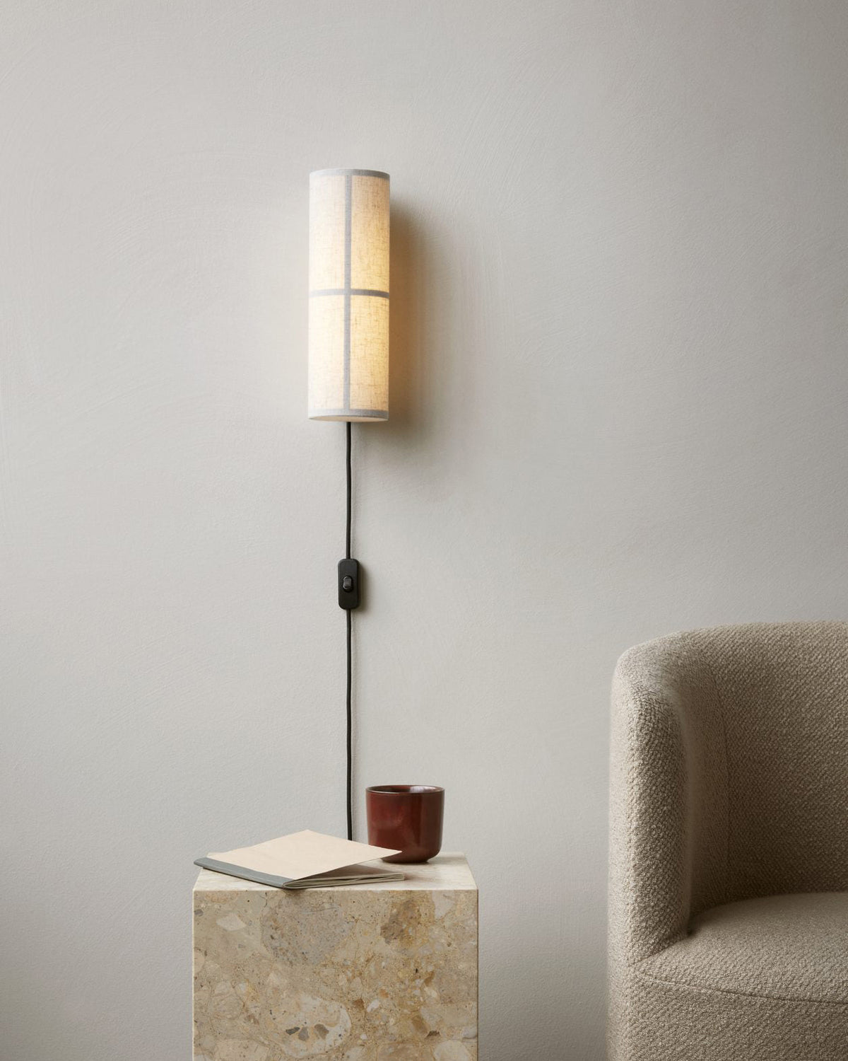 A white Clérachic fabric wall light above a stone side table with a book and a red cup, a boucle armchair at the right.