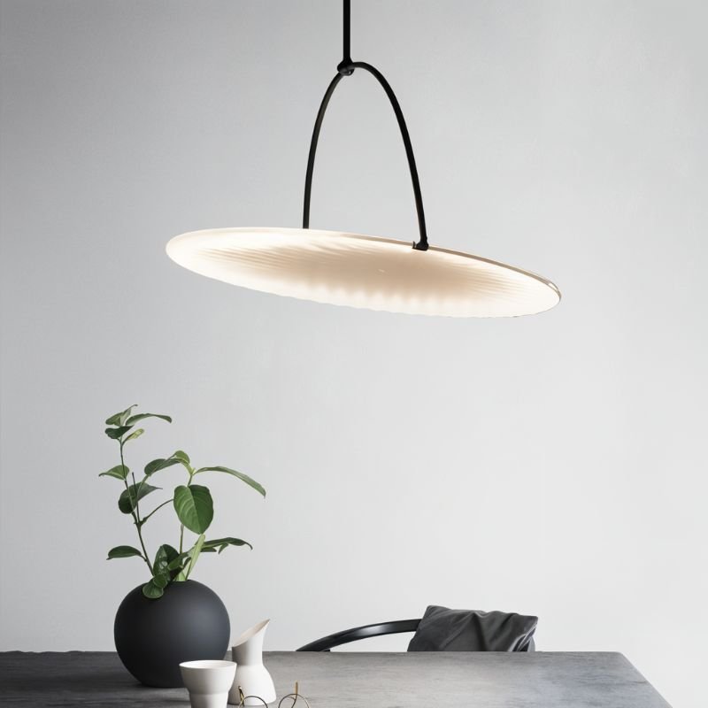 Clérachic pendant with a tilted pleated white disc on a slim black arched hanger above a grey table with a plant and vase.