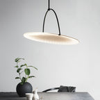 Clérachic pendant with a tilted pleated white disc on a slim black arched hanger above a grey table with a plant and vase.