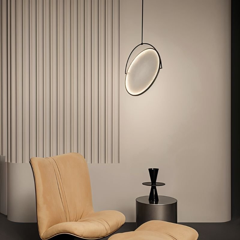 Clérachic black-framed round pendant with an LED-edged disc, tilted on a ceiling rod beside a tan armchair and fluted wall.