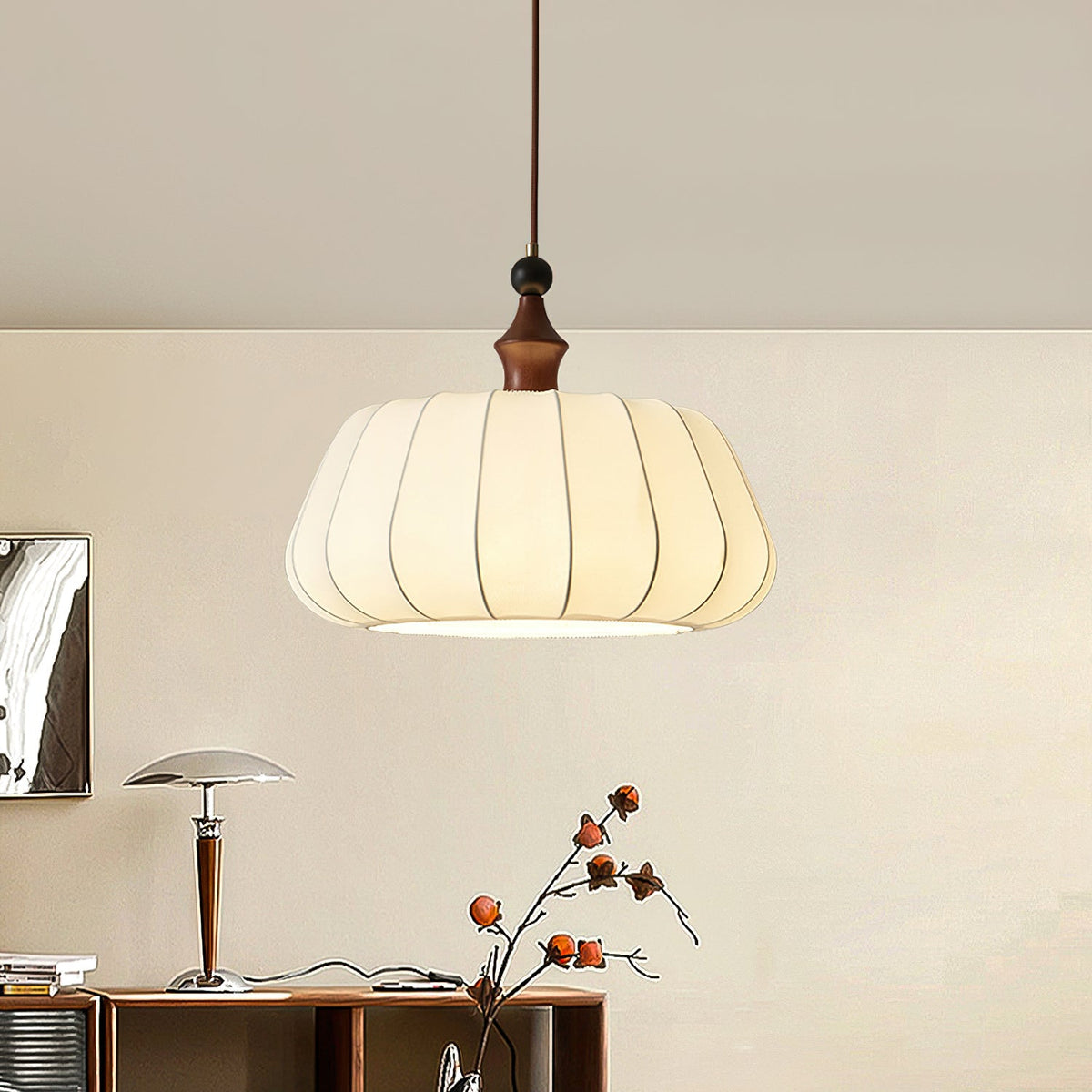 A lit Clérachic fabric dome pendant hangs above a wooden sideboard with a mushroom-shaped table lamp and a branch with orange fruit.