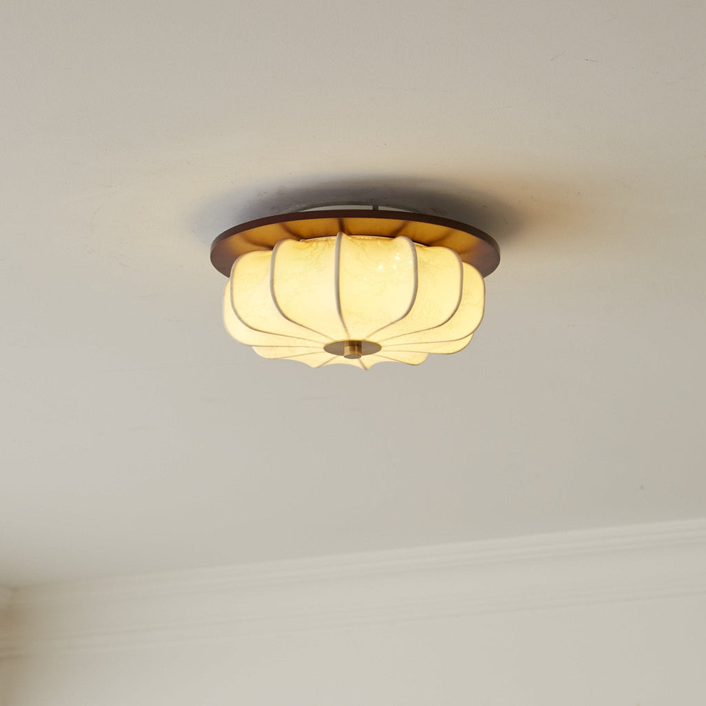 A lit Clérachic fabric ceiling light with a walnut rim and brass cap, above a ceiling cornice, glowing a warm cream.