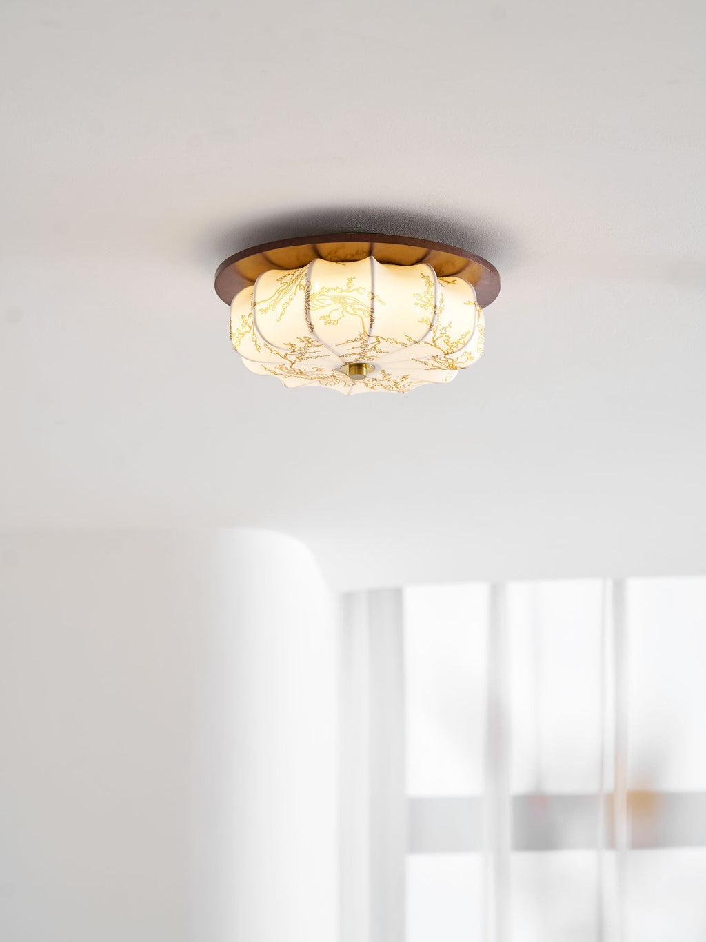 A lit Clérachic ceiling light with a walnut rim and floral-etched fabric shade, near a white wall and sheer curtain.