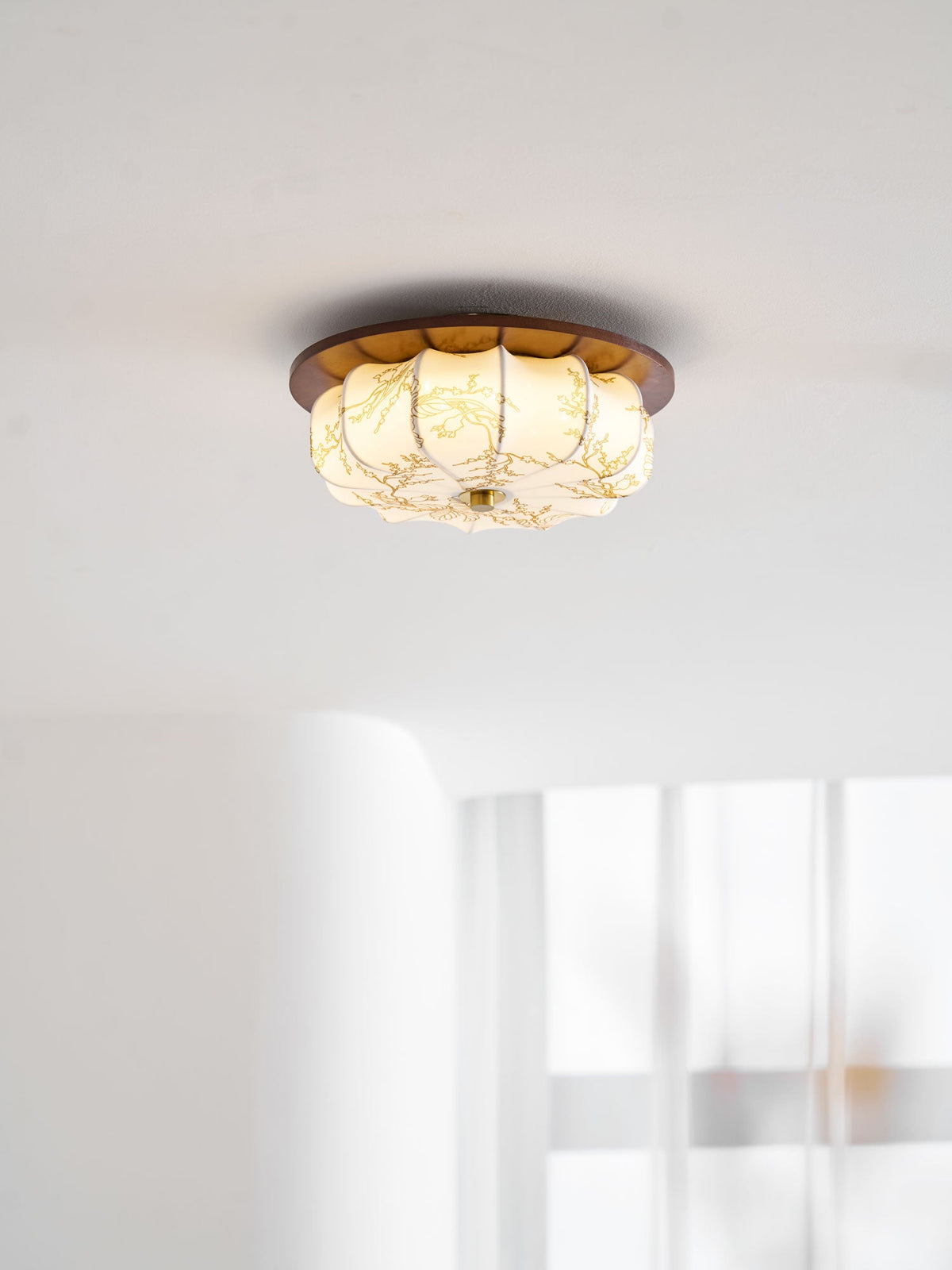 A lit Clérachic ceiling light with a walnut rim and floral-etched fabric shade, near a white wall and sheer curtain.