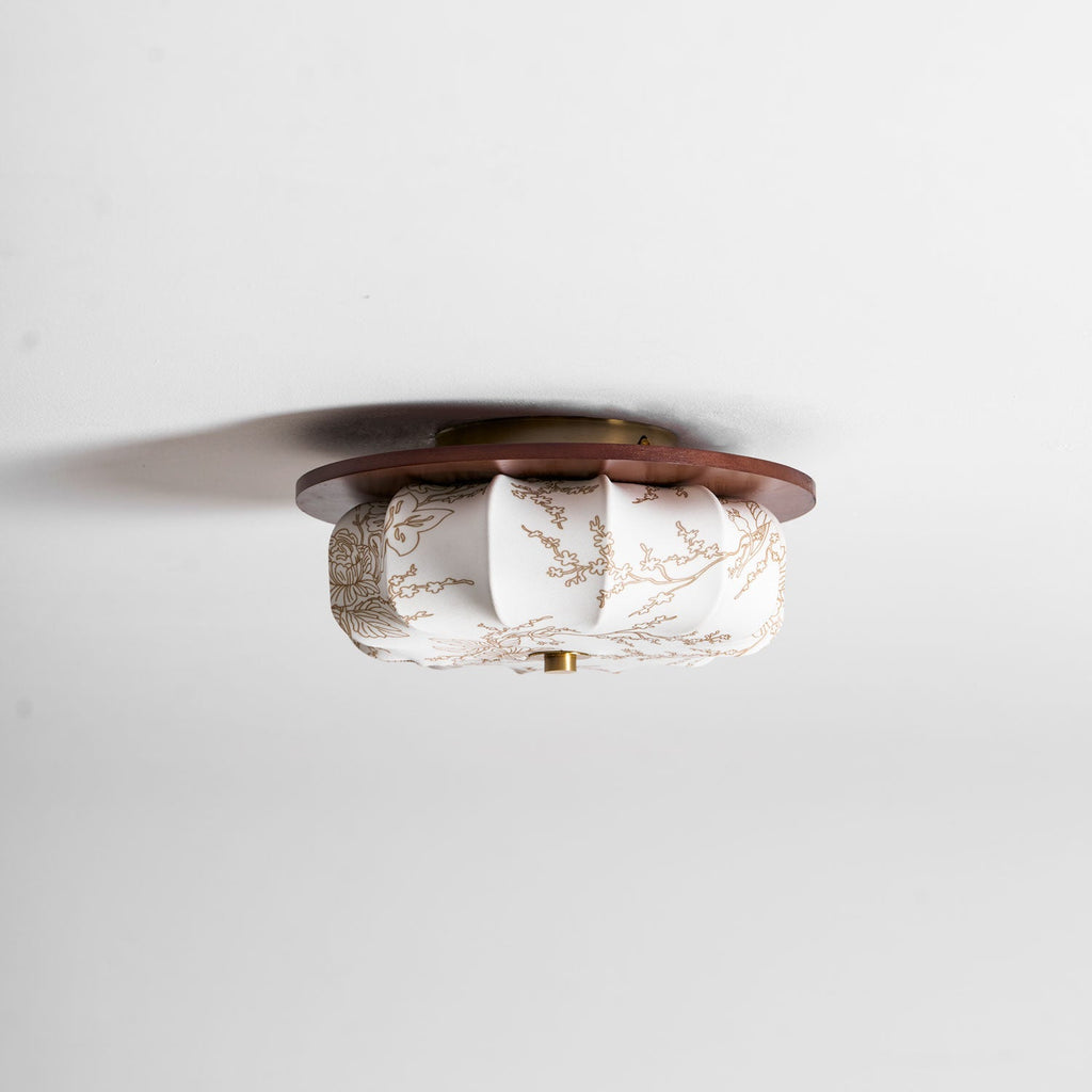 A Clérachic ceiling light seen from the side, showing the walnut disc, brass cap and floral-etched fabric shade.