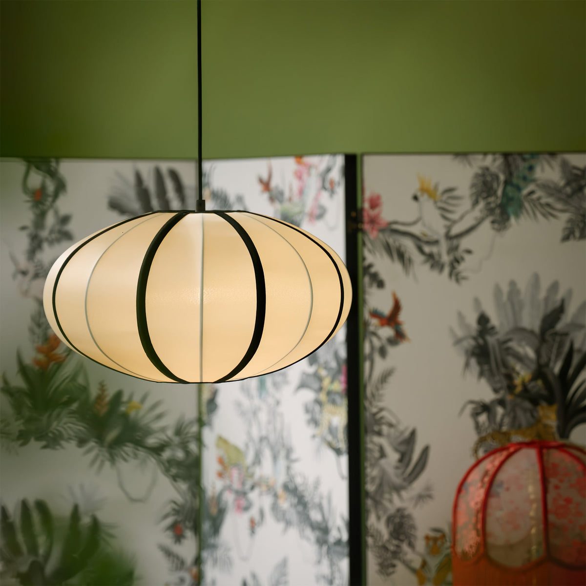 A lit oval fabric pendant with black ribs hangs in front of a botanical screen and an olive-green wall, a Clérachic fabric pendant.