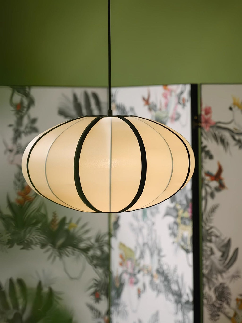 A softly glowing fabric pendant with black ribbing hangs in front of a floral folding screen under an olive-green ceiling, from Clérachic.