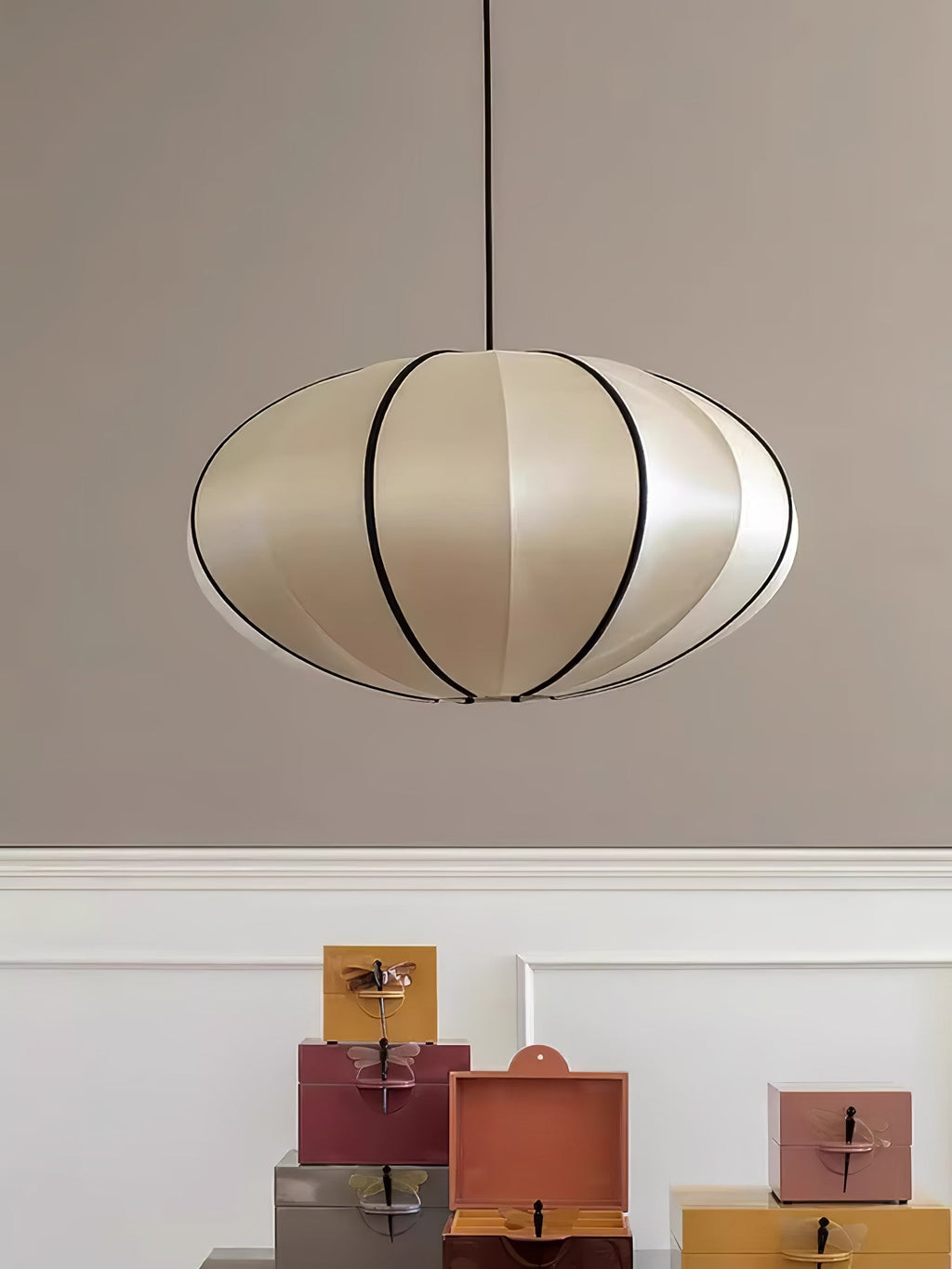A flattened fabric pendant in a pale silver-cream shade with black ribs hangs against a taupe wall above a row of small boxes, from Clérachic.