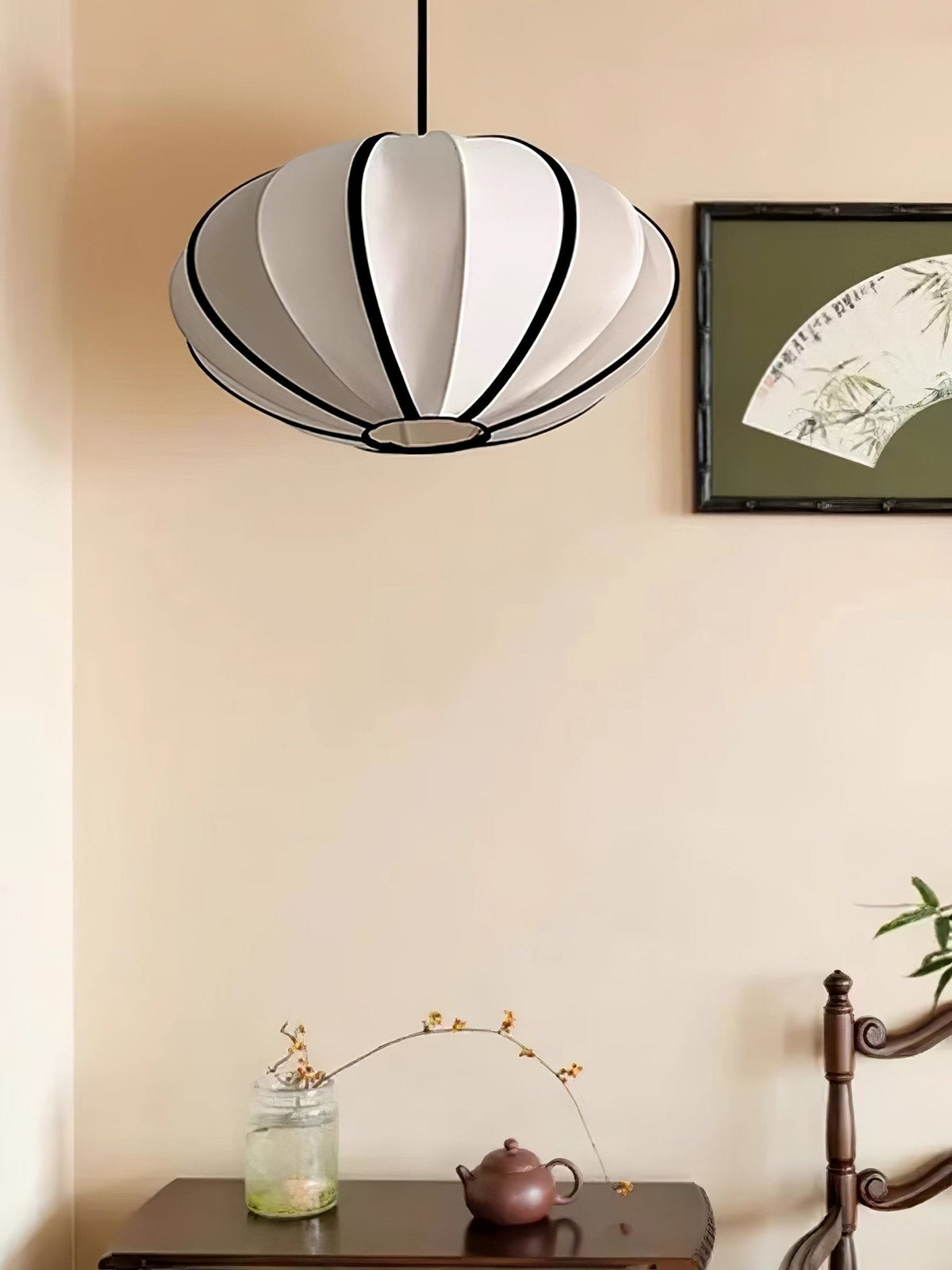 A white fabric pendant with black ribs hangs against a peach wall beside a framed fan painting and a small console, from Clérachic.