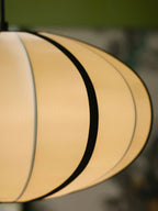 Close view of the pale fabric panels and one black rib on this Clérachic pendant, glowing warmly against an olive backdrop.
