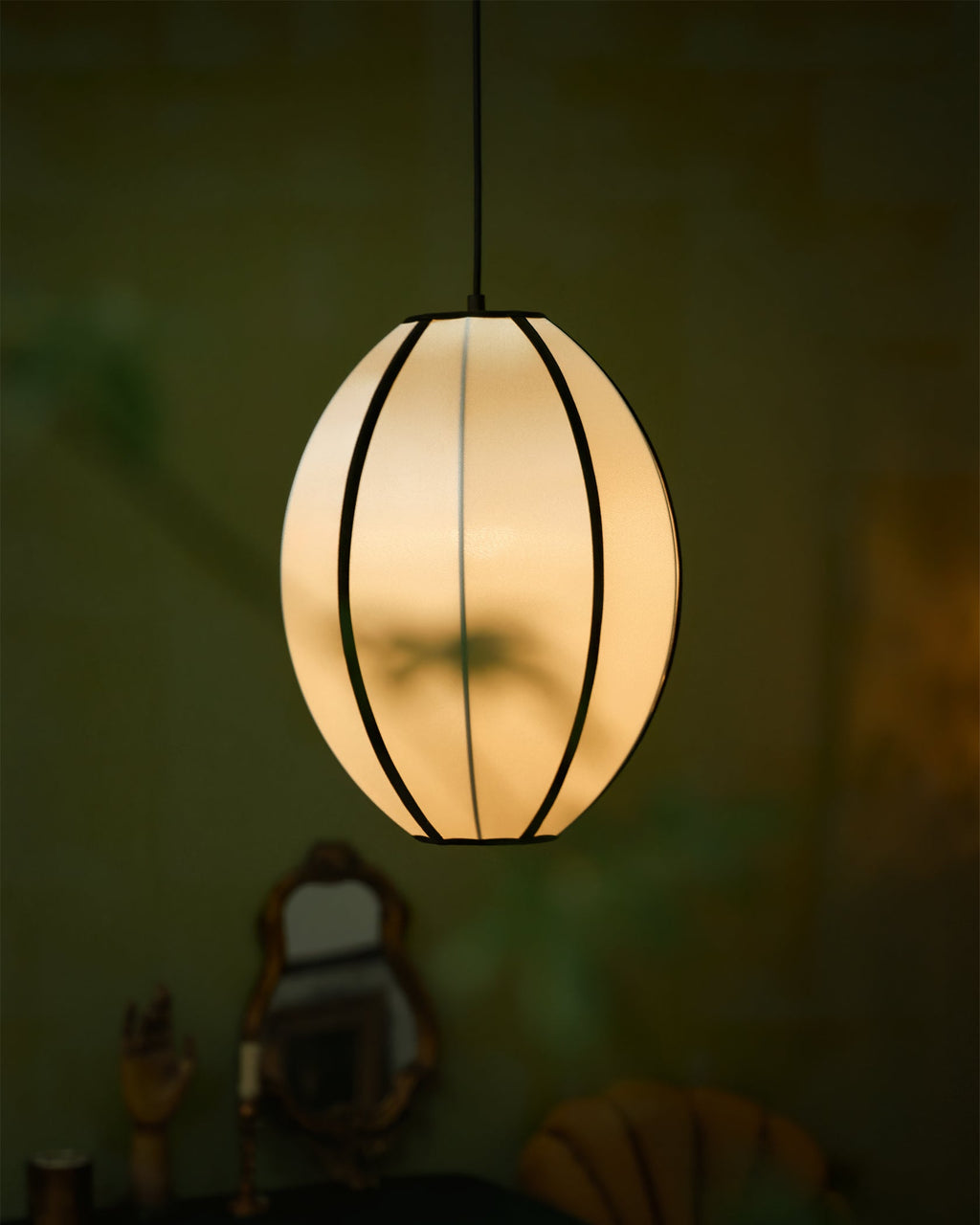 A small egg-shaped fabric pendant with black ribs glows warm in a dim green room with gilt mirrors, from Clérachic.