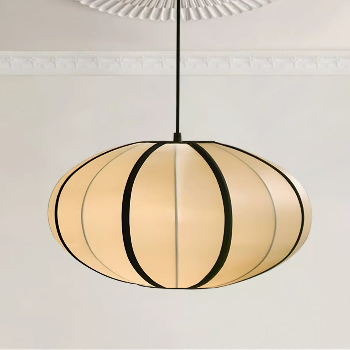 A wide, flattened fabric pendant with bold black ribs hangs from a black cord below a decorative plaster ceiling rose, from Clérachic.