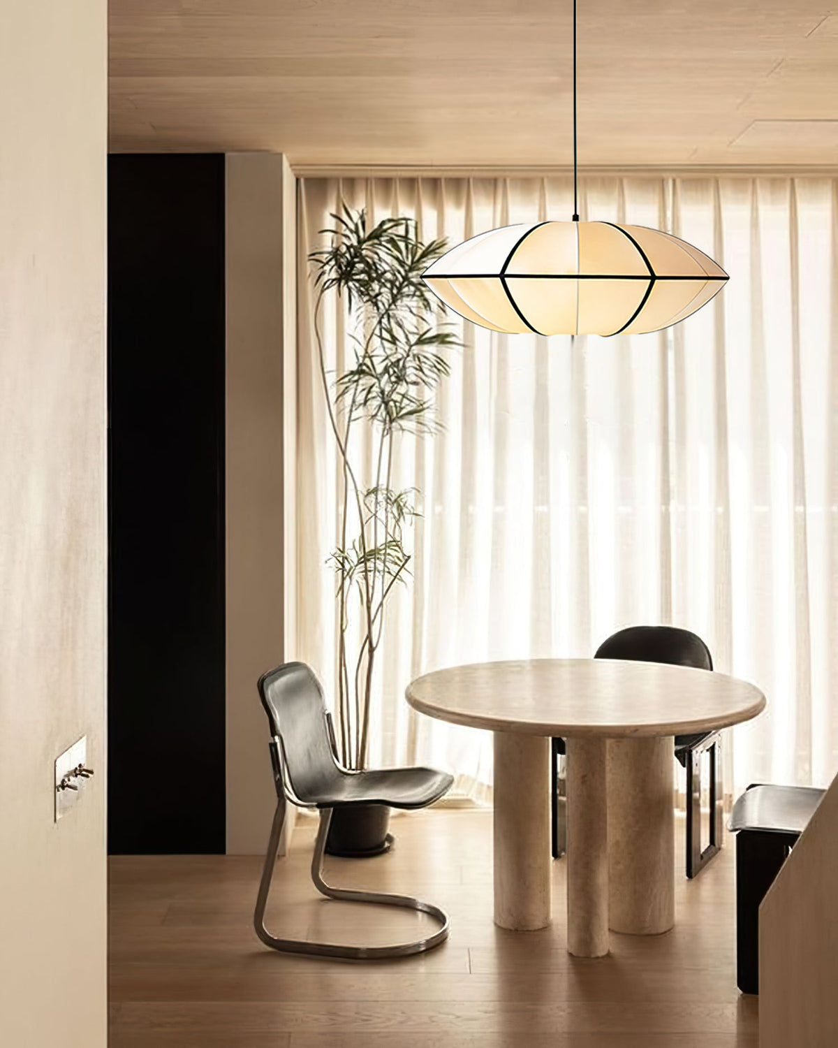 A flat fabric pendant with black ribs hangs beside sheer curtains over a round stone table and a chrome chair, from Clérachic.