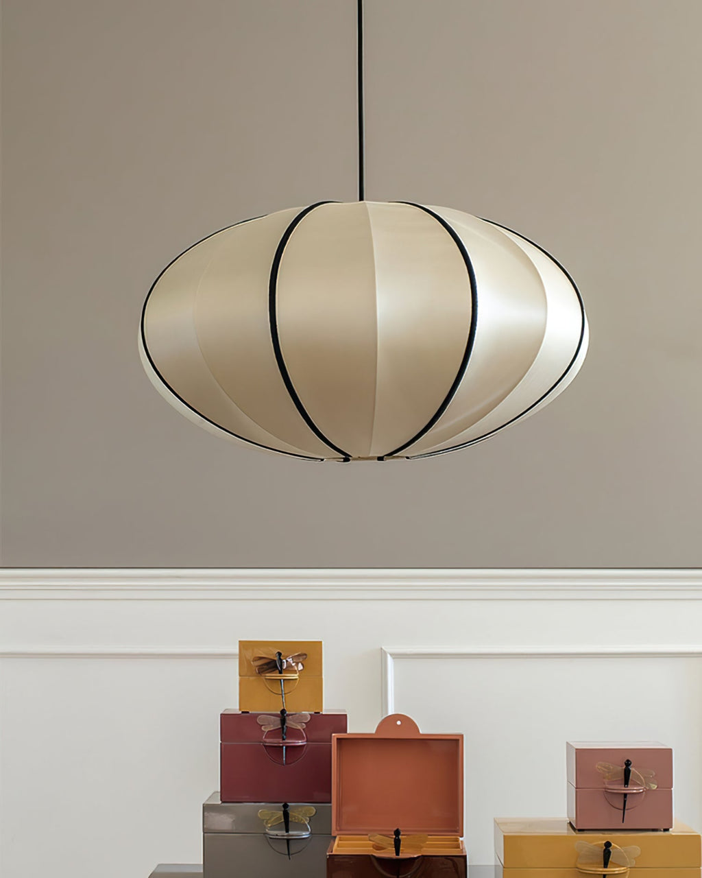 A flattened, pale fabric pendant with black ribs hangs against a grey wall above a white panel and a stack of coloured boxes, from Clérachic.