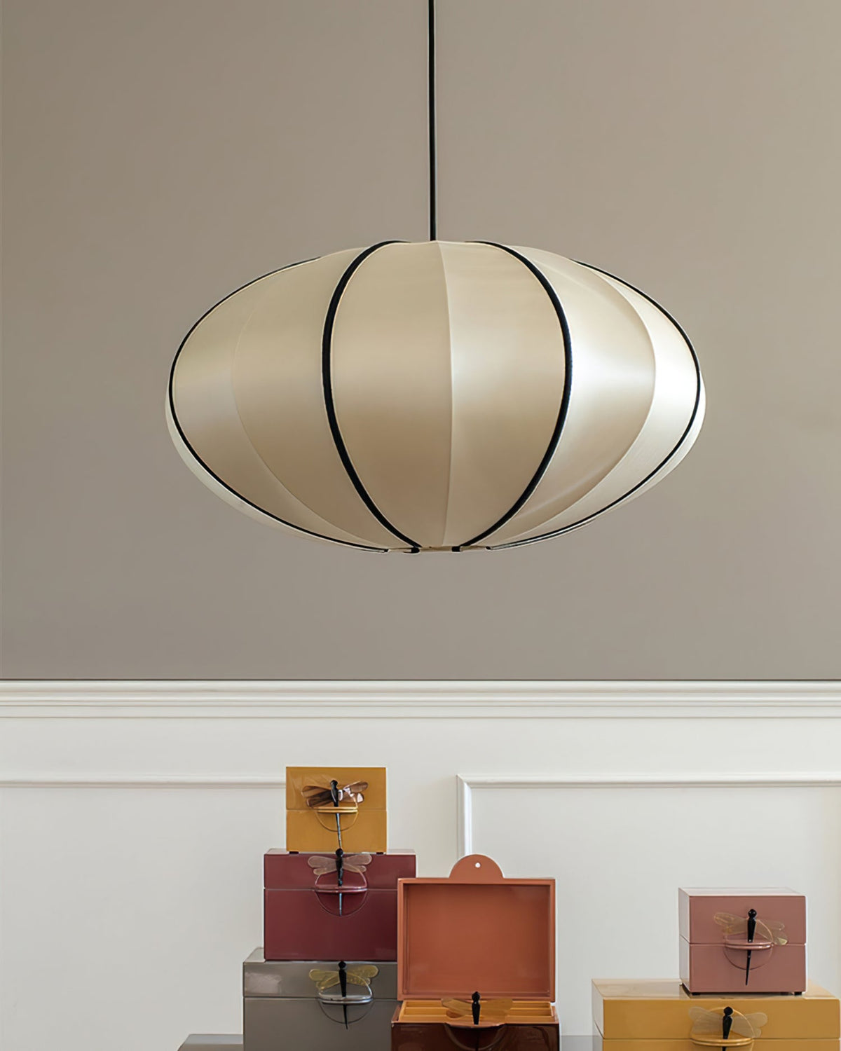 A flattened, pale fabric pendant with black ribs hangs against a grey wall above a white panel and a stack of coloured boxes, from Clérachic.