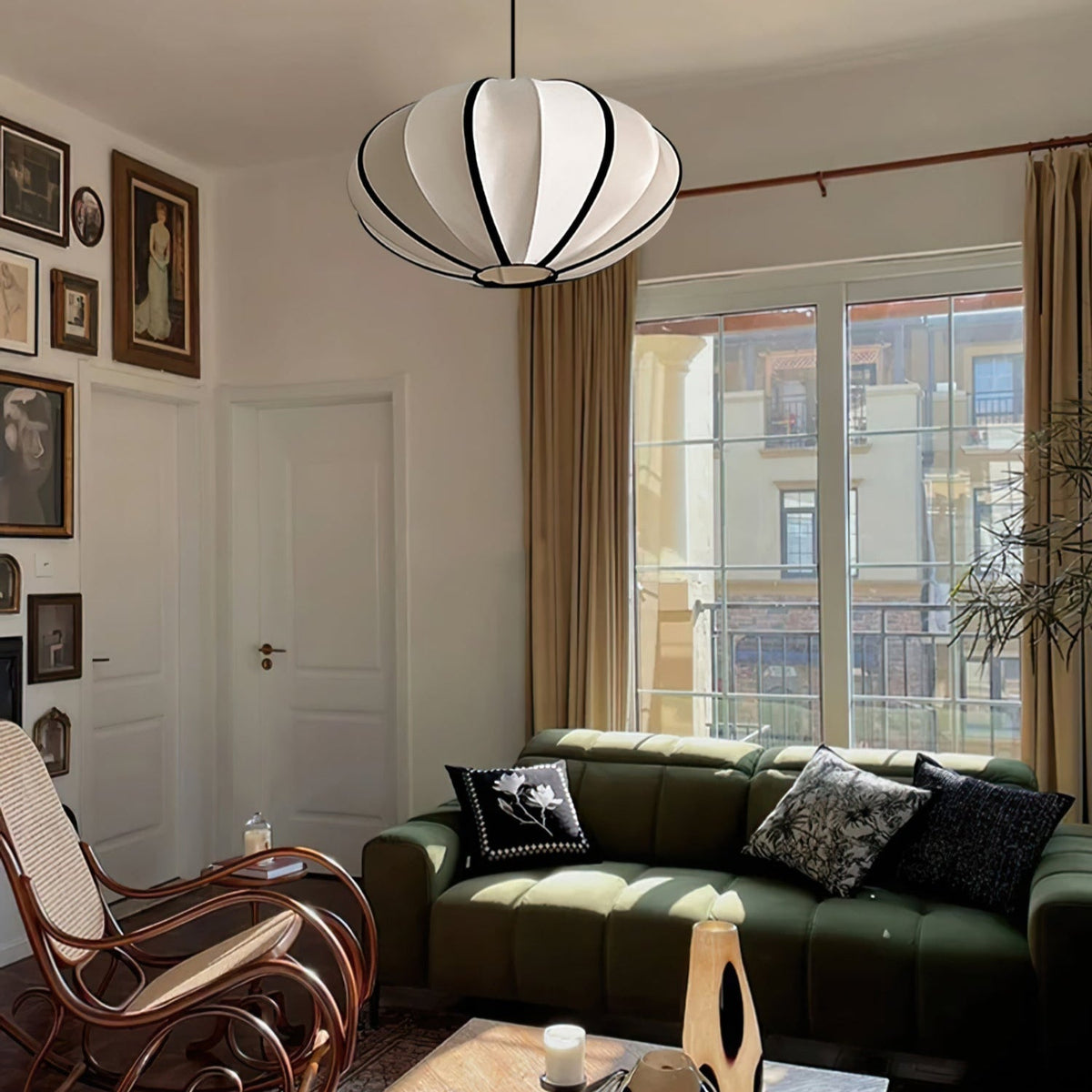 A white fabric pendant with black ribs hangs in a living room with a green sofa, a cane rocking chair and framed pictures, from Clérachic.