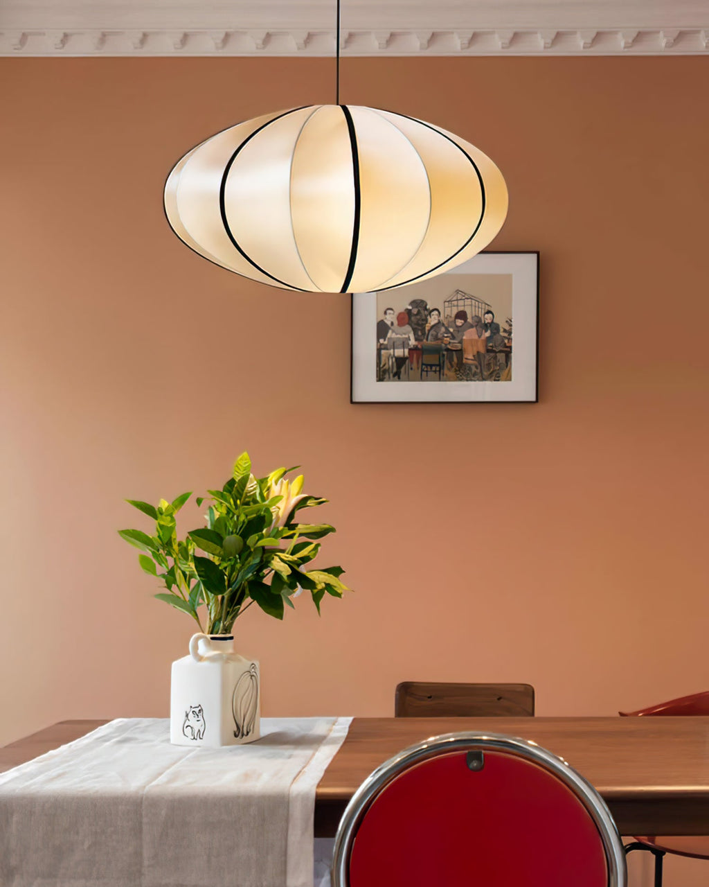 A fabric pendant with black ribbing glows against a terracotta wall, above a dining table set with a vase of leaves, from Clérachic.