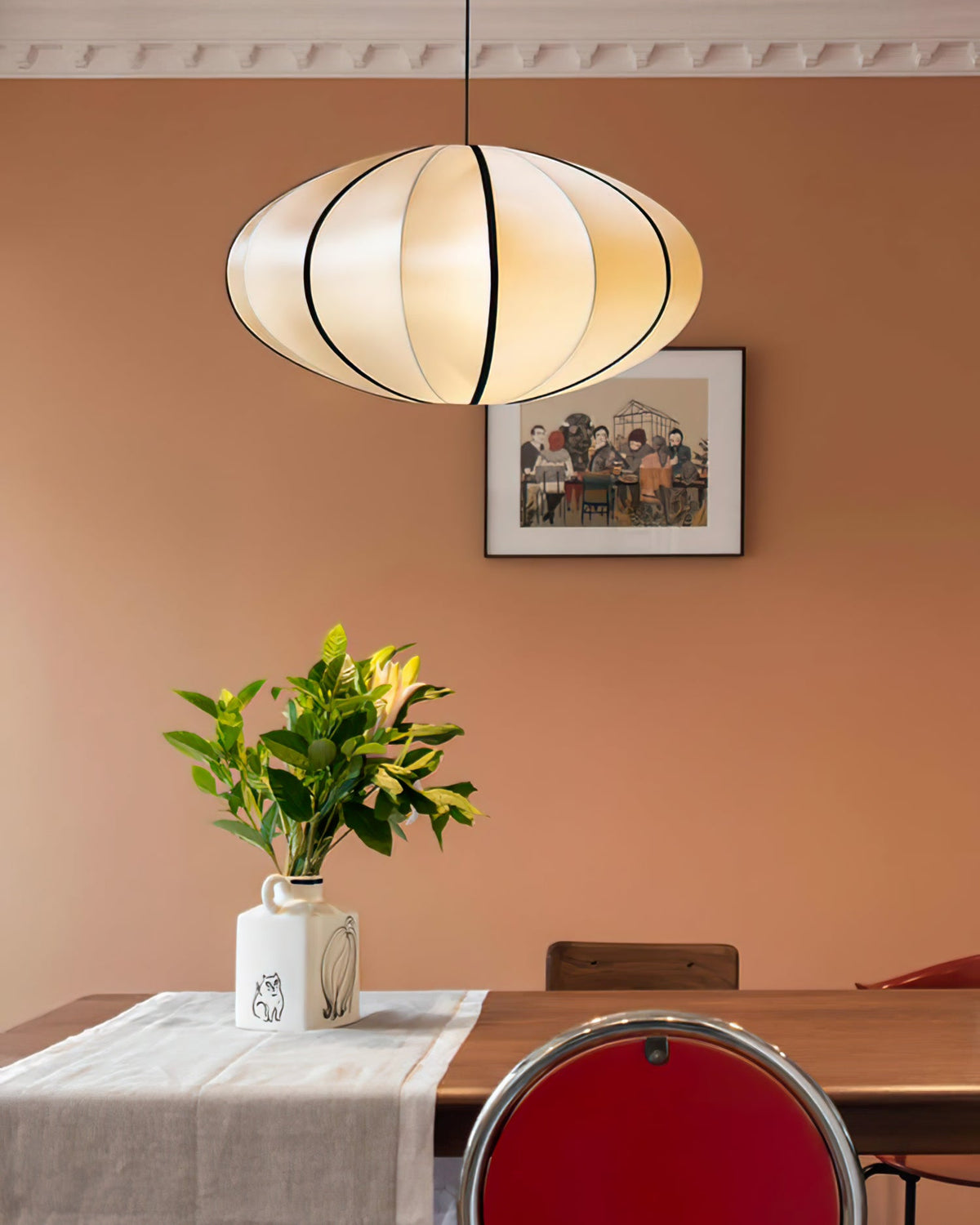 A fabric pendant with black ribbing glows against a terracotta wall, above a dining table set with a vase of leaves, from Clérachic.