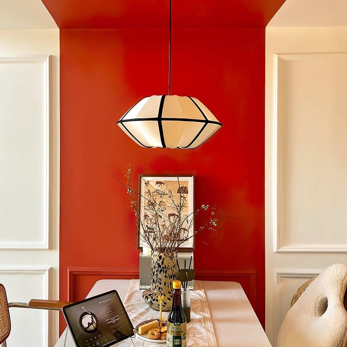 A white fabric pendant with a faceted, angular shape and black ribs hangs against a red wall above a dining table, from Clérachic.
