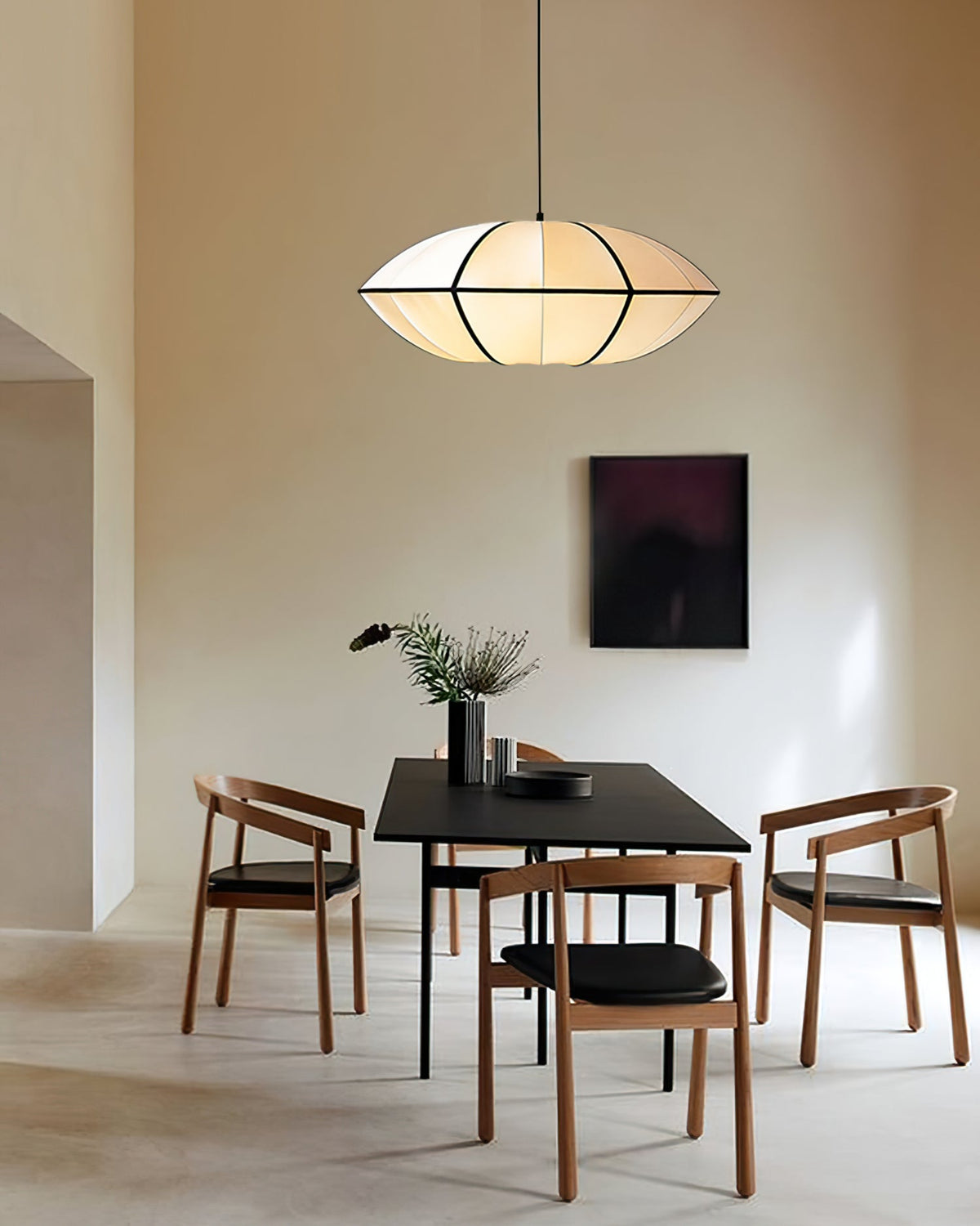 A white fabric pendant with a pointed rim hangs above a black table with four wooden chairs in a plain cream room, from Clérachic.