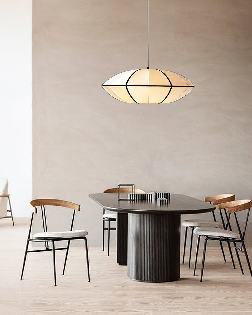 A white fabric pendant with a sharp edge hangs above a dark oval table with wooden chairs against a plaster wall, from Clérachic.