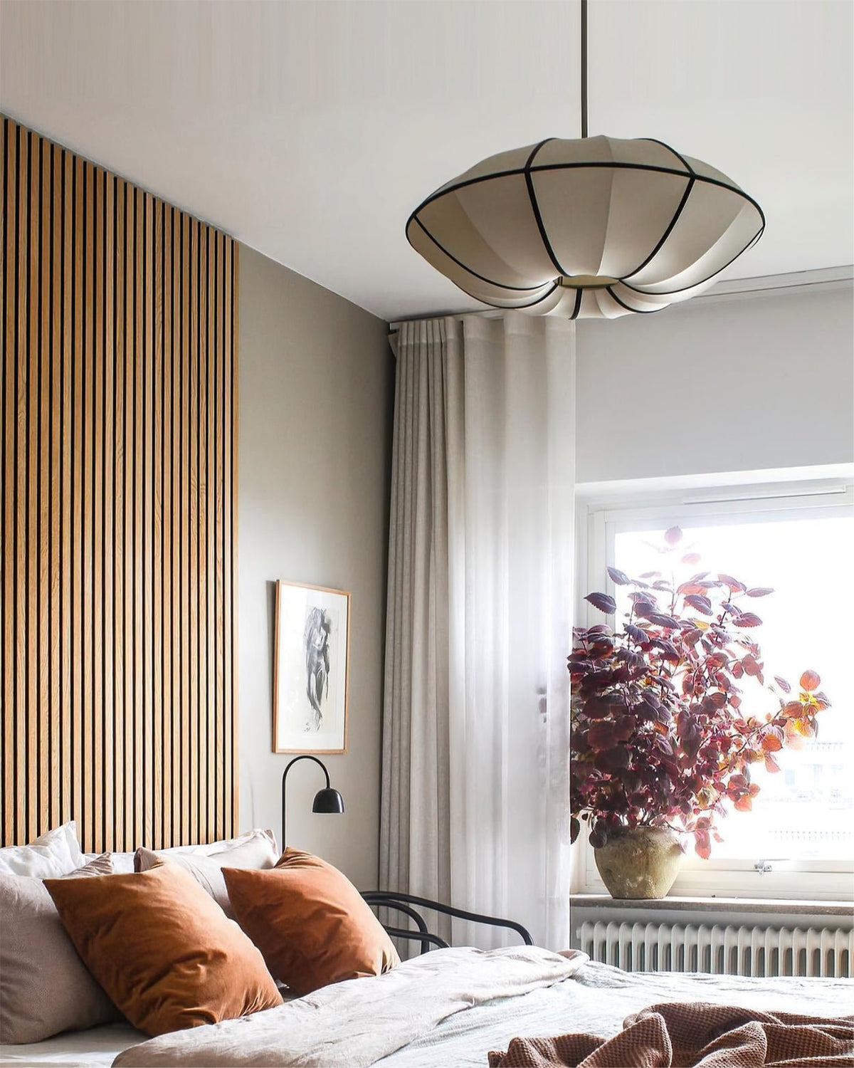 A white, scalloped fabric pendant with black ribs hangs above a bed with rust cushions, beside a timber-slat wall, from Clérachic.