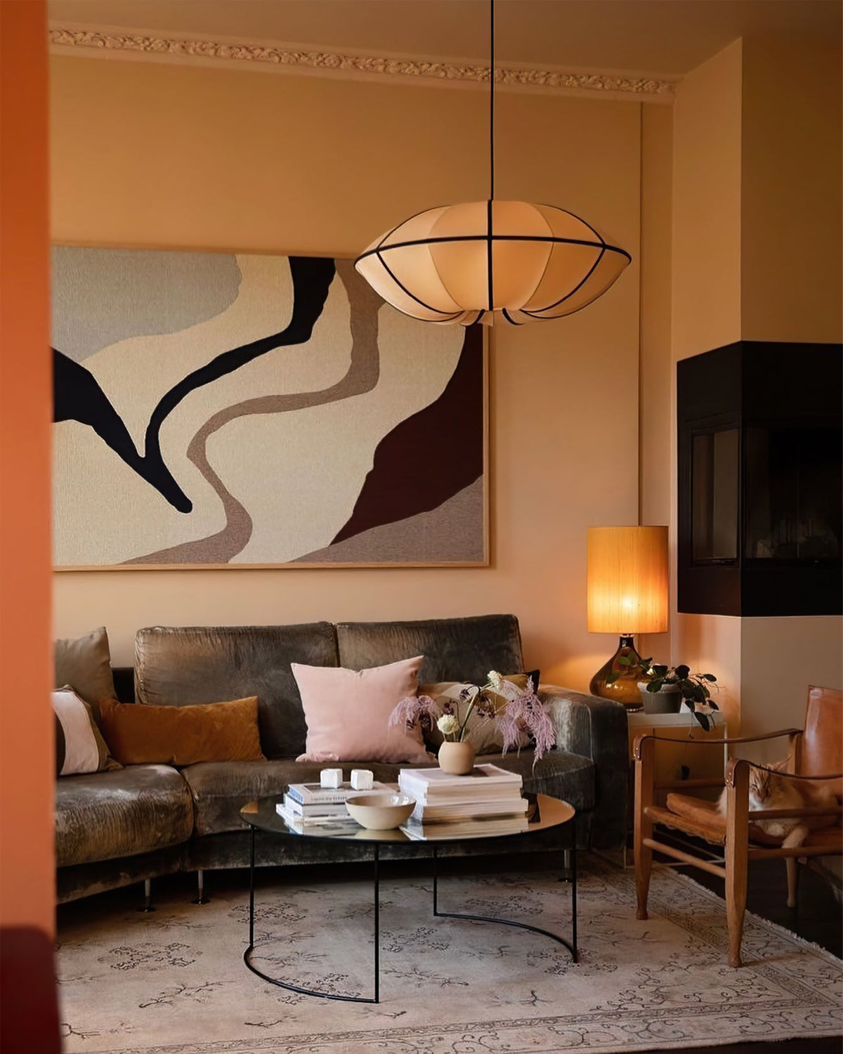 A fabric pendant with black ribs glows warm above a velvet sofa and round glass coffee table in a peach-toned living room, from Clérachic.
