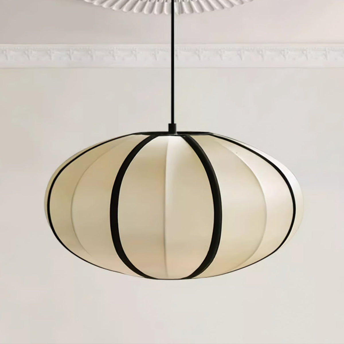 A flattened fabric pendant with bold black ribs hangs from a black cord, a moulded ceiling above, a wide Clérachic shade.