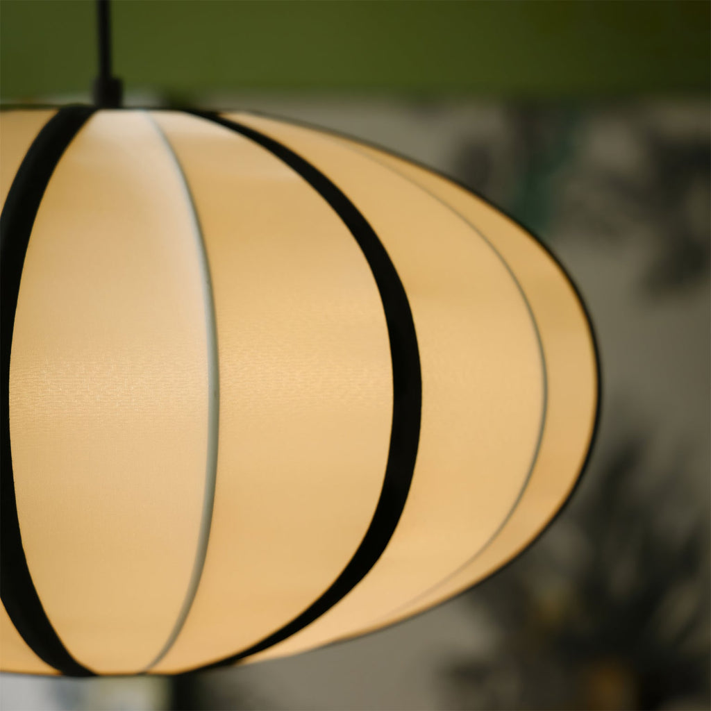 Close view of the glowing cream fabric and black ribs of this Clérachic pendant, with soft green leaves out of focus behind.