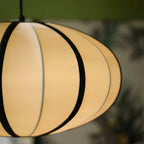 Close view of the glowing cream fabric and black ribs of this Clérachic pendant, with soft green leaves out of focus behind.