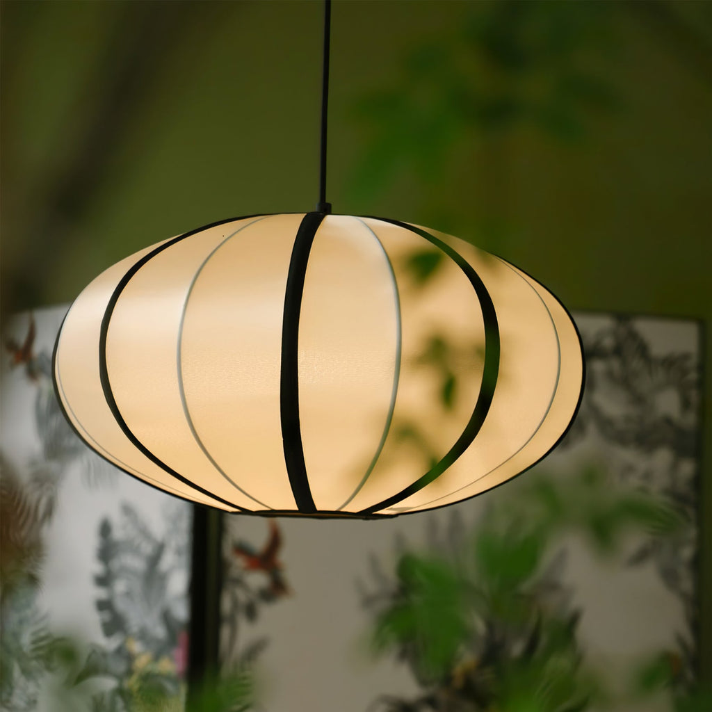 A lit oval fabric pendant with black ribs hangs against blurred green leaves and a floral screen, from Clérachic.