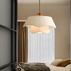 This Clérachic pendant hangs in a bedroom, its two ruffled fabric layers lit against a pale curtain beside an unmade bed.