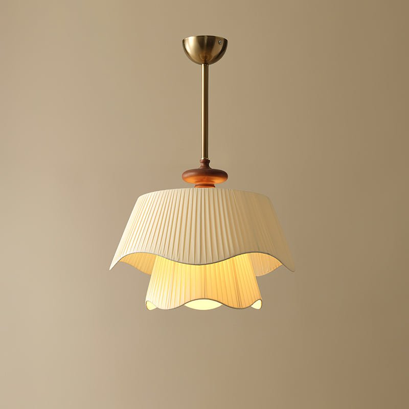 Lit Clérachic pendant on a plain beige wall, its inner fabric layer glowing a warm yellow beneath the wavy outer shade.