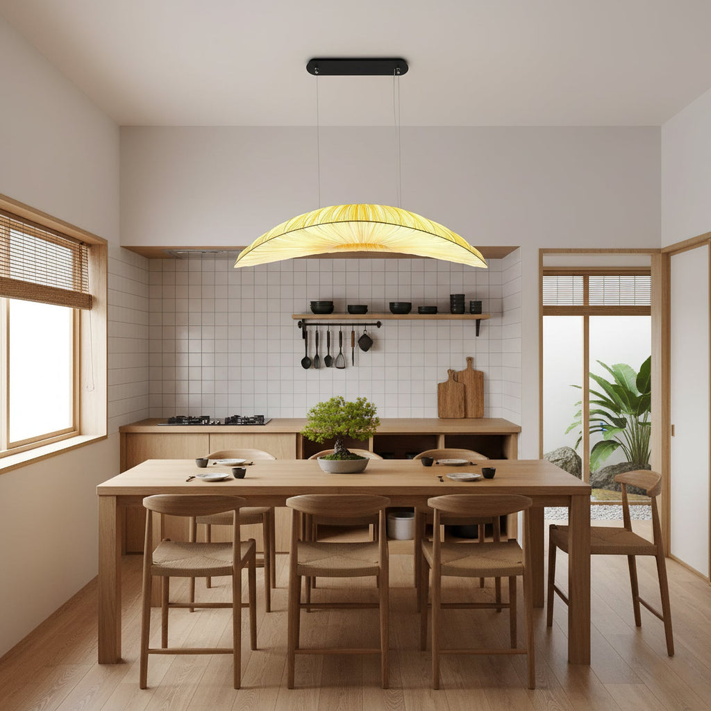 A Clérachic leaf-shaped pendant glows yellow above a wooden dining table with woven chairs, in a kitchen with white tile and open shelves.