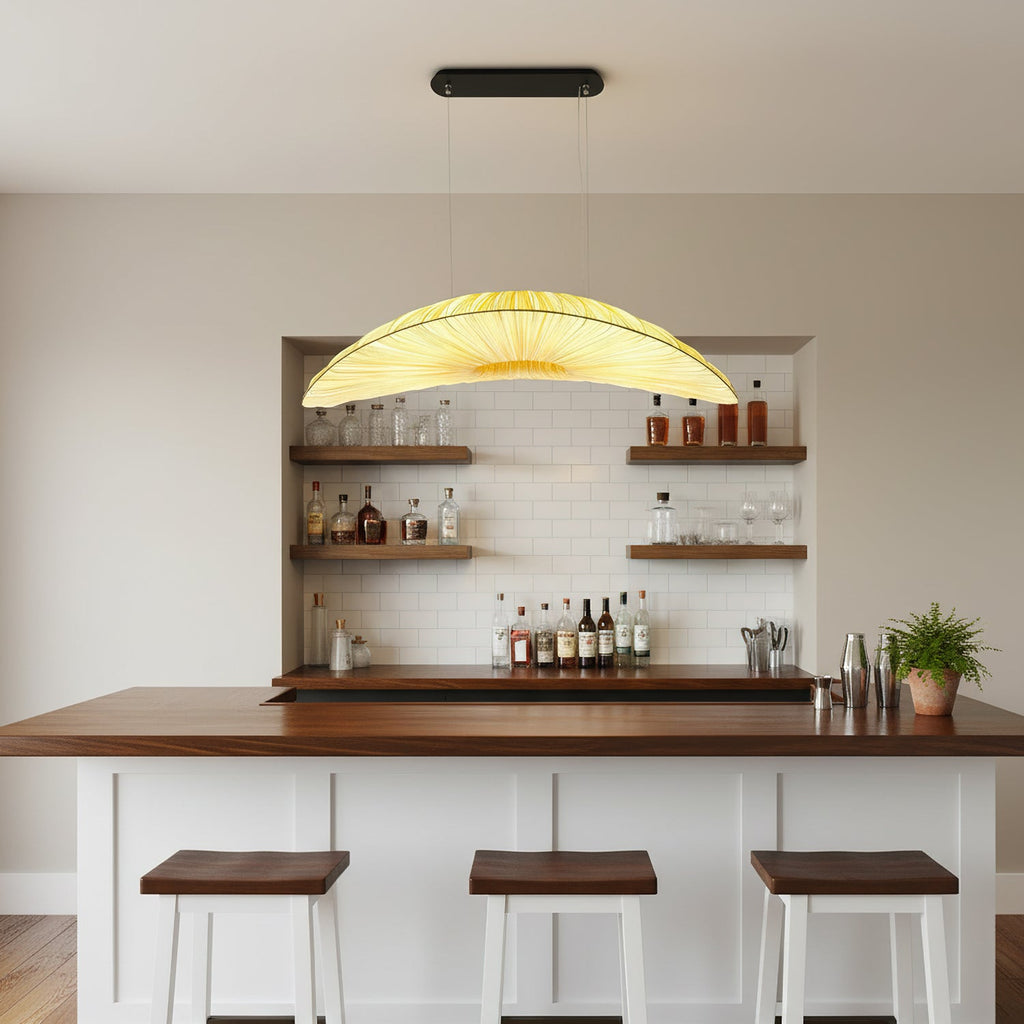 Over a bar counter with dark wood stools, a Clérachic pendant with a pleated fabric shade hangs in front of shelves of bottles.