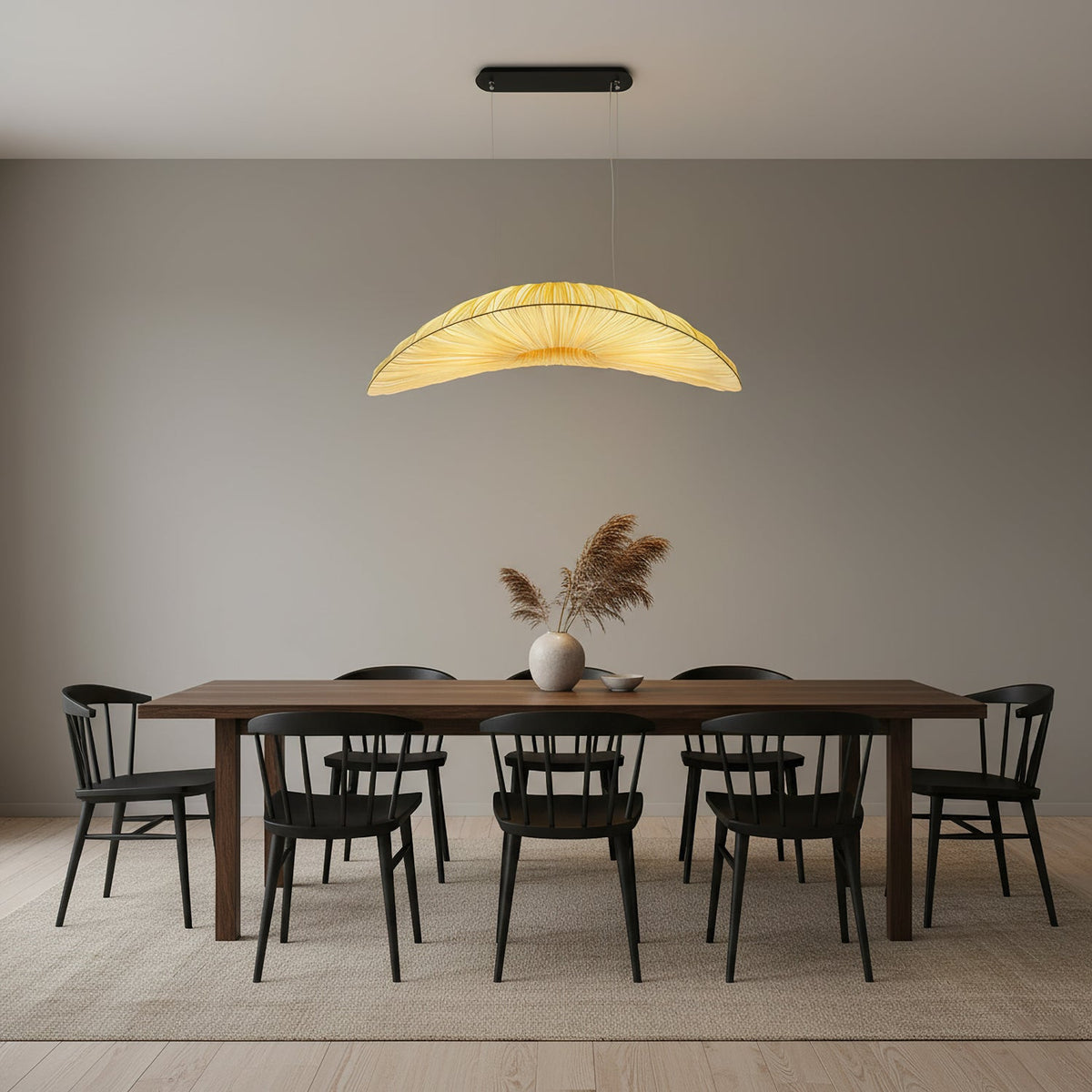 A Clérachic pendant with a glowing leaf-shaped shade hangs over a long dark dining table and black chairs, in a calm grey room.