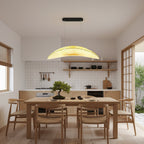 A Clérachic pendant with a pleated, glowing shade hangs over a dining table with wishbone chairs, in front of a white tiled kitchen.
