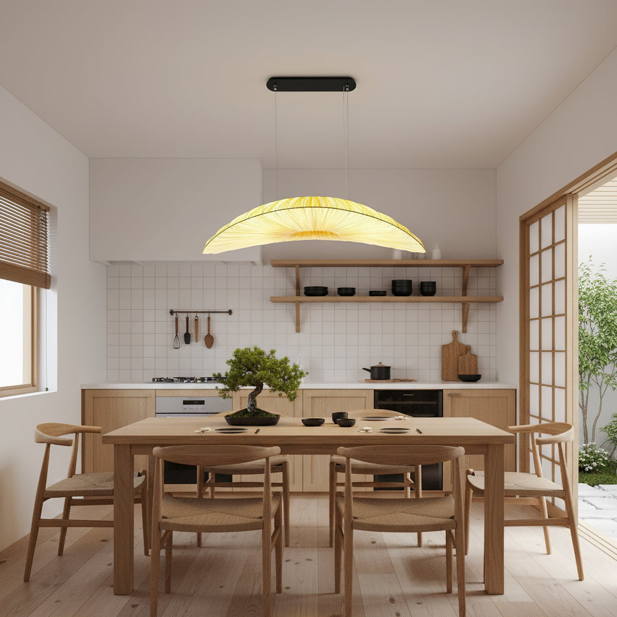 A Clérachic pendant with a pleated, glowing shade hangs over a dining table with wishbone chairs, in front of a white tiled kitchen.