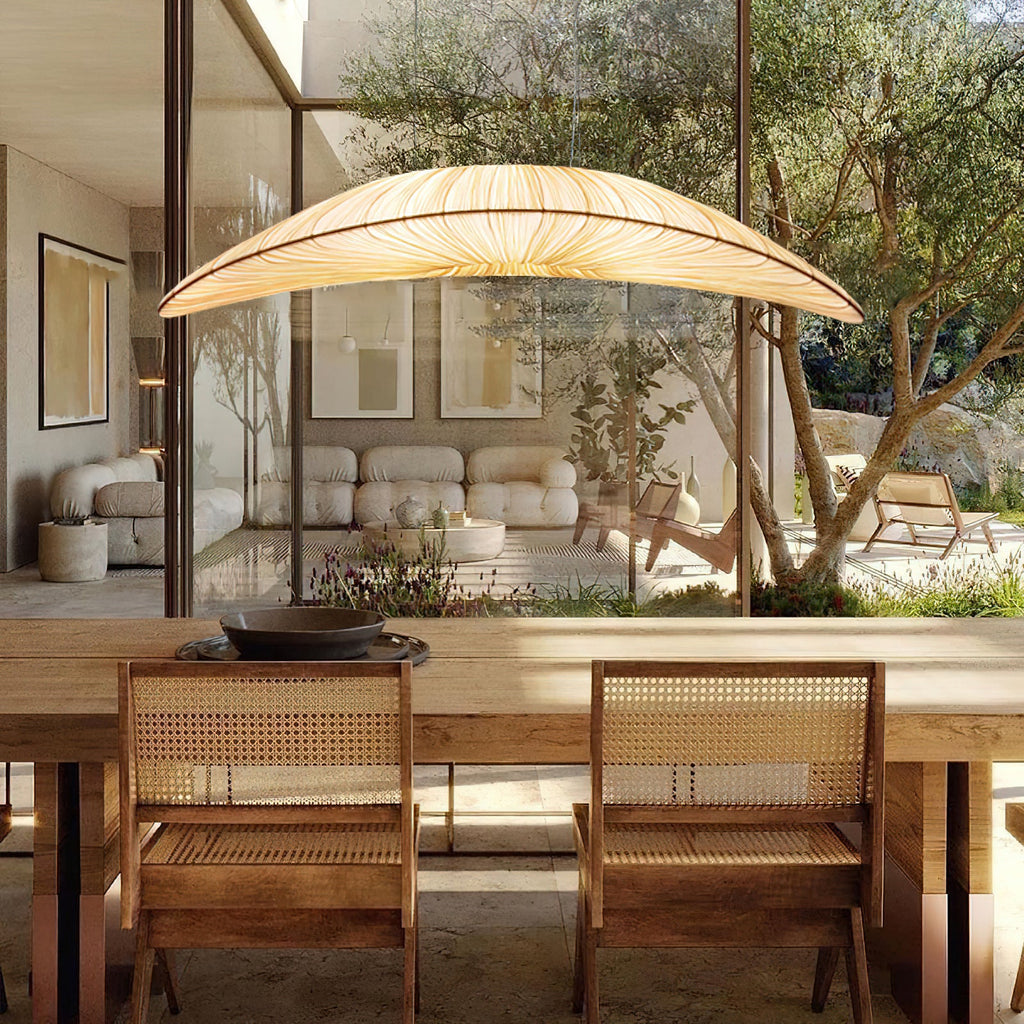 A wide Clérachic pendant with a cream pleated shade hangs above a dining table with cane chairs, with an olive tree outside the glass.