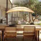 A wide Clérachic pendant with a cream pleated shade hangs above a dining table with cane chairs, with an olive tree outside the glass.