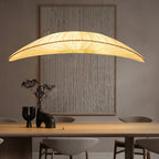 Over a wooden table with a dark vase, a Clérachic pendant with a long pleated shade hangs in front of a ridged wall panel.