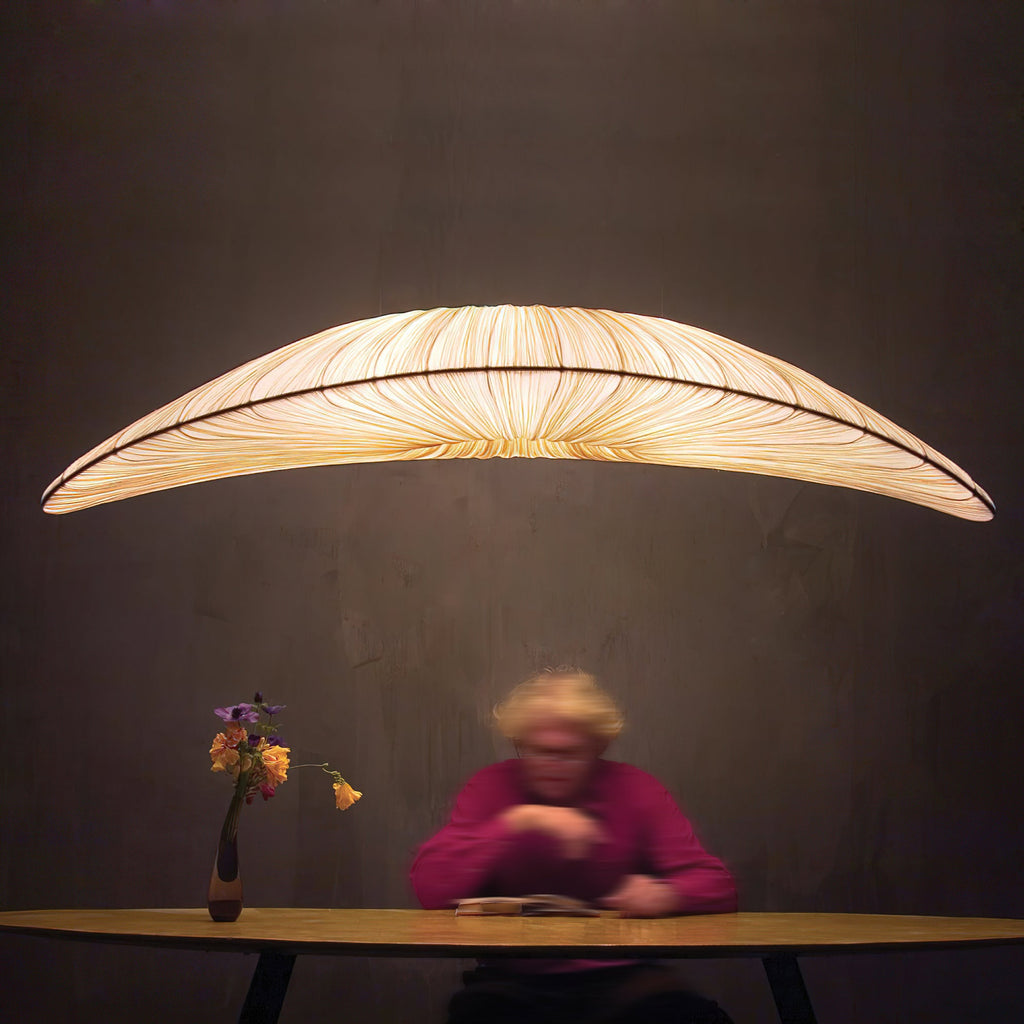 A Clérachic pendant glows warm over a round table in a dark room, where a person in a magenta top sits with a small vase of flowers.