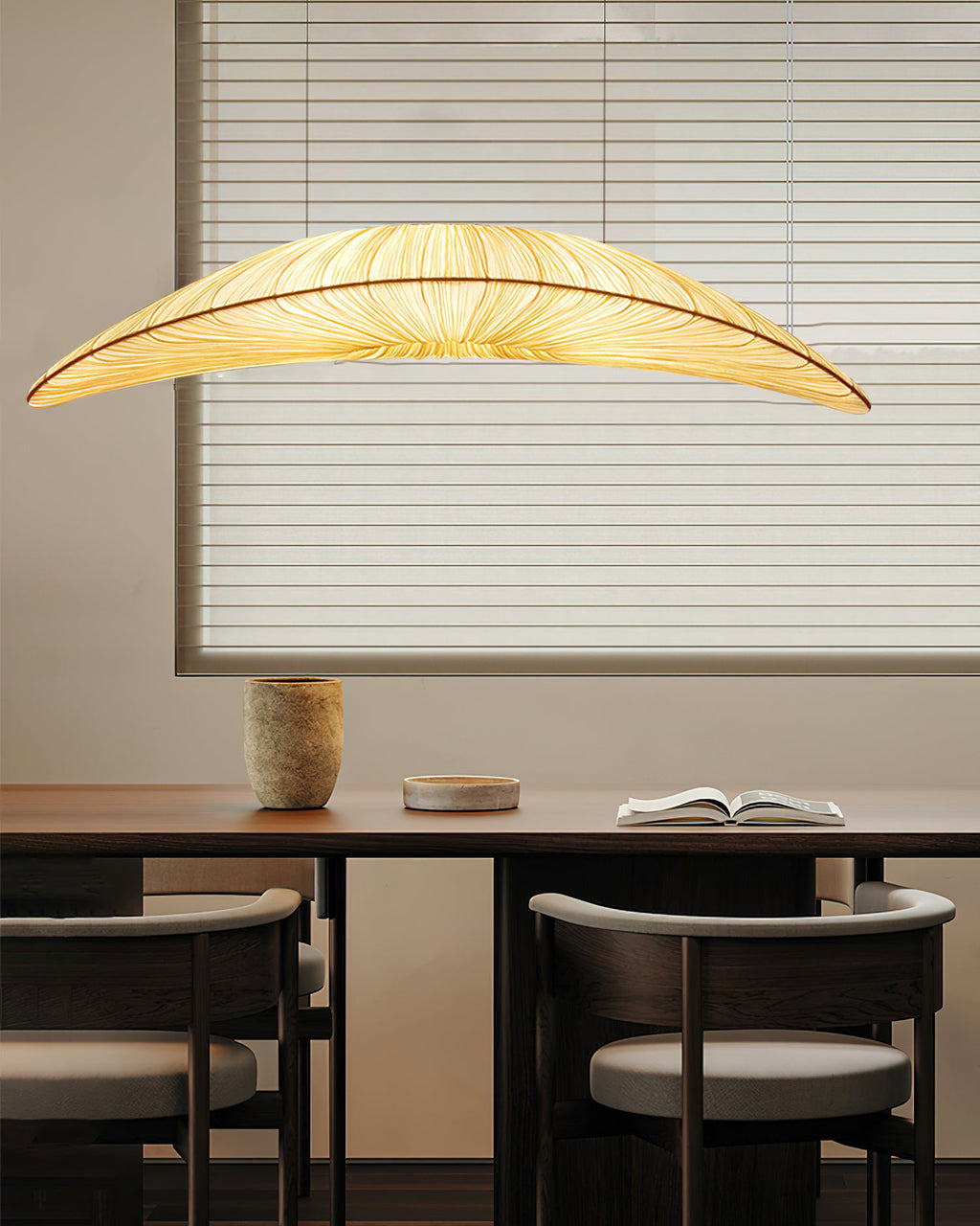 Above a dark dining table with rounded chairs, a Clérachic pendant with a long leaf-shaped shade hangs in front of a blind.