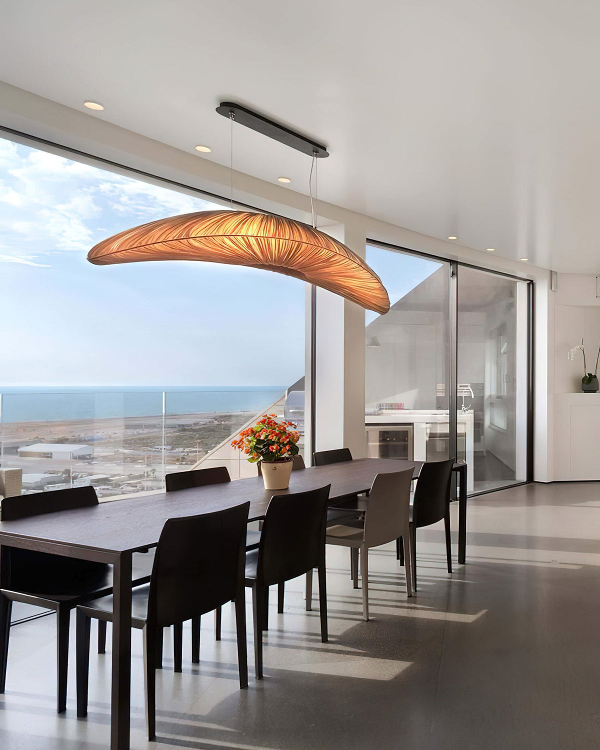 A Clérachic pendant in an amber glow hangs over a long dark dining table, with a wide glass wall and the sea in view.