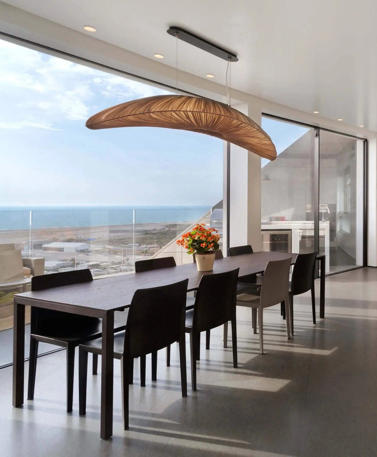 A Clérachic pendant with a darker, amber-brown shade hangs in front of a glass wall that looks out over a coastline.
