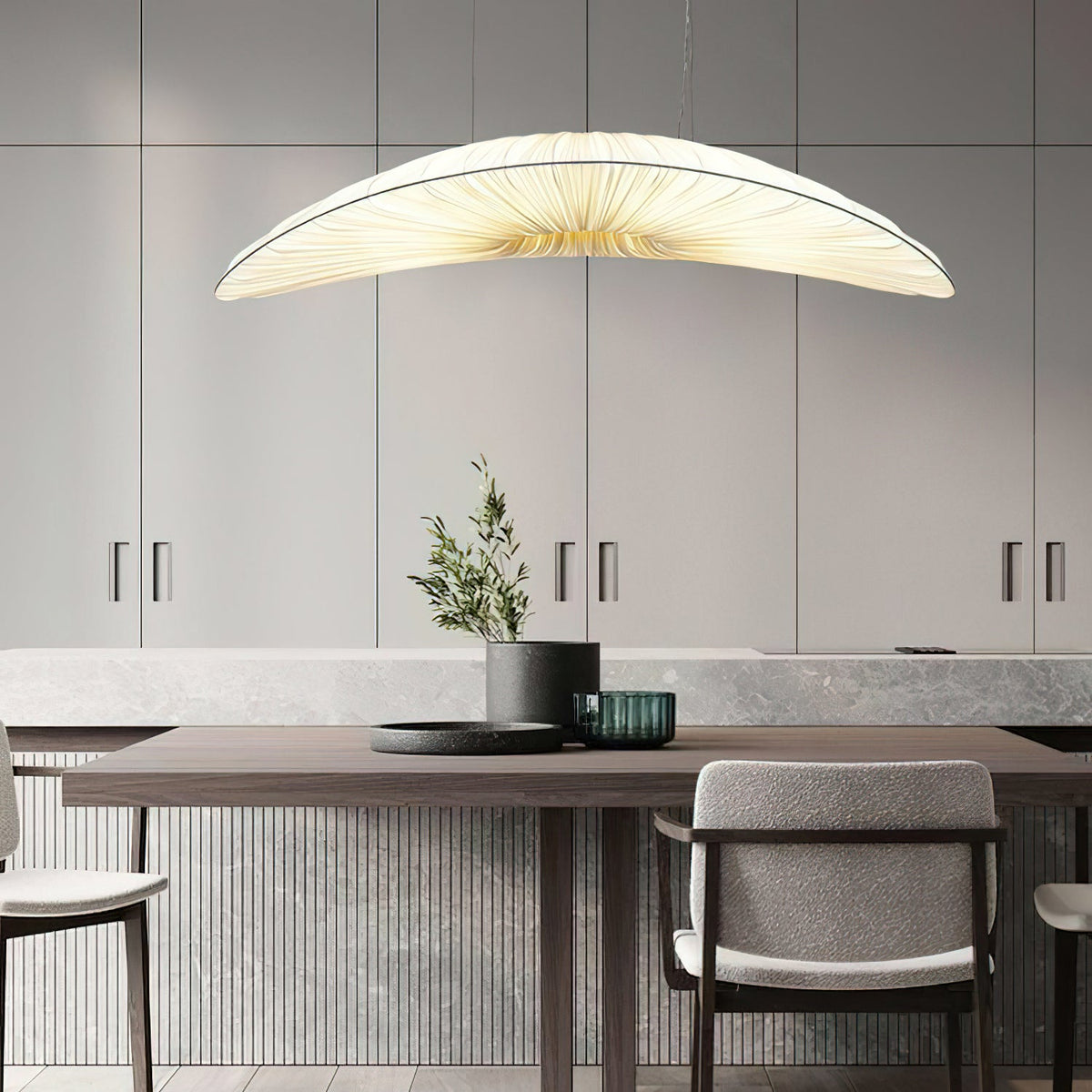 A Clérachic pendant with a cream pleated shade hangs over a table with a small olive plant, in front of grey cabinets.