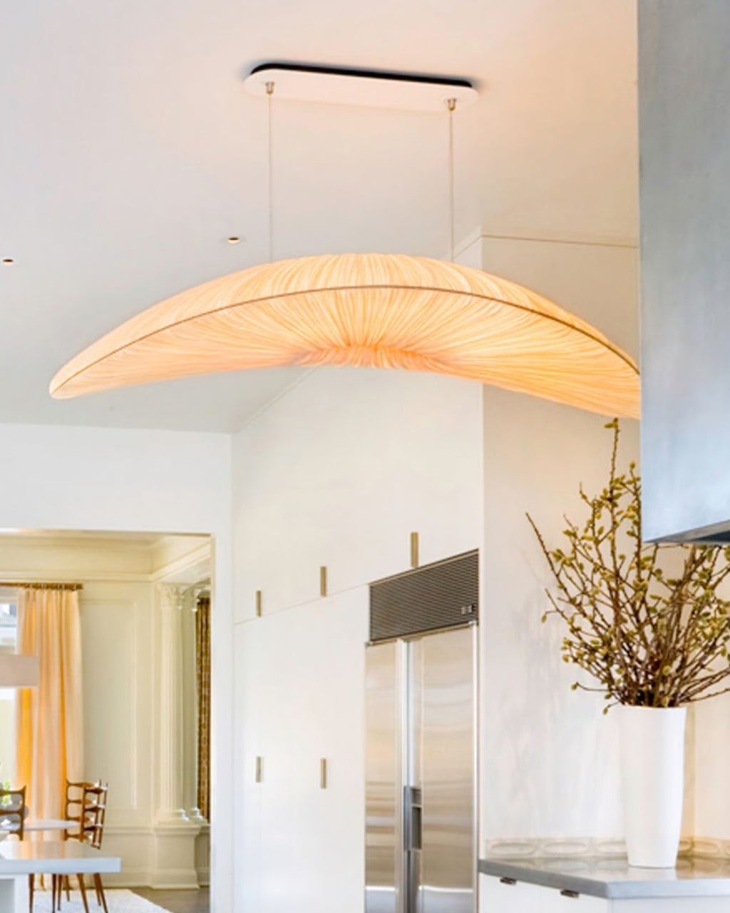 A Clérachic pendant with a peach-toned pleated shade hangs from a black plate, in a bright white room with a steel fridge.