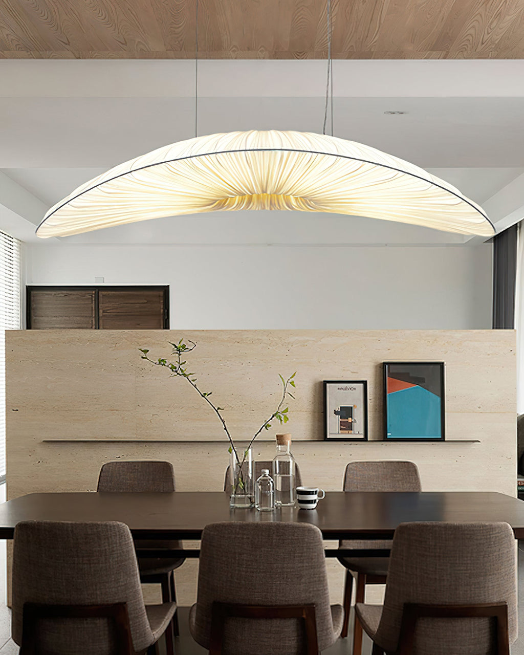 A Clérachic pendant with a pale pleated shade hangs above a dining table with brown chairs, in front of a stone-look shelf.