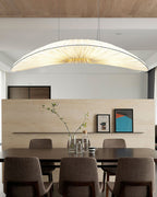 A Clérachic pendant with a pale pleated shade hangs above a dining table with brown chairs, in front of a stone-look shelf.