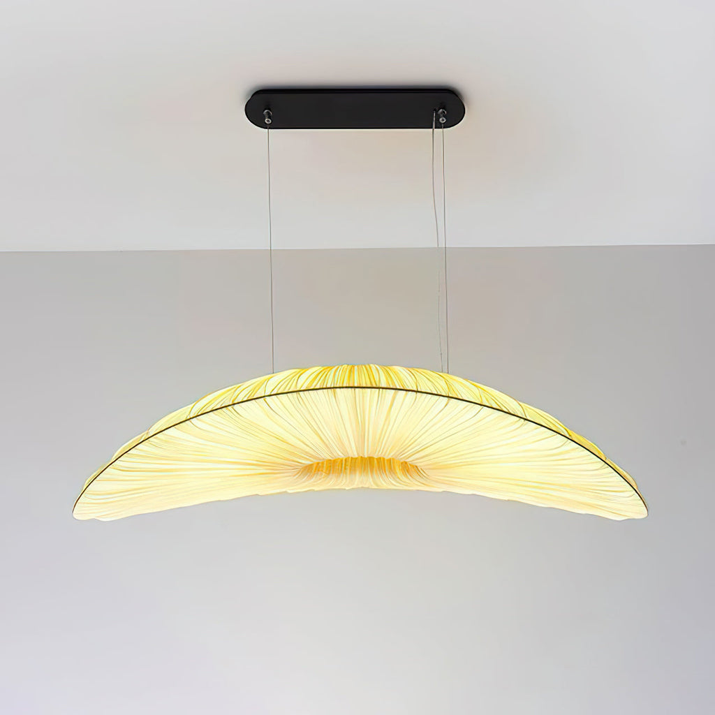 A Clérachic pendant with a black oval ceiling plate and a pale pleated shade that glows yellow in the middle, against a plain wall.